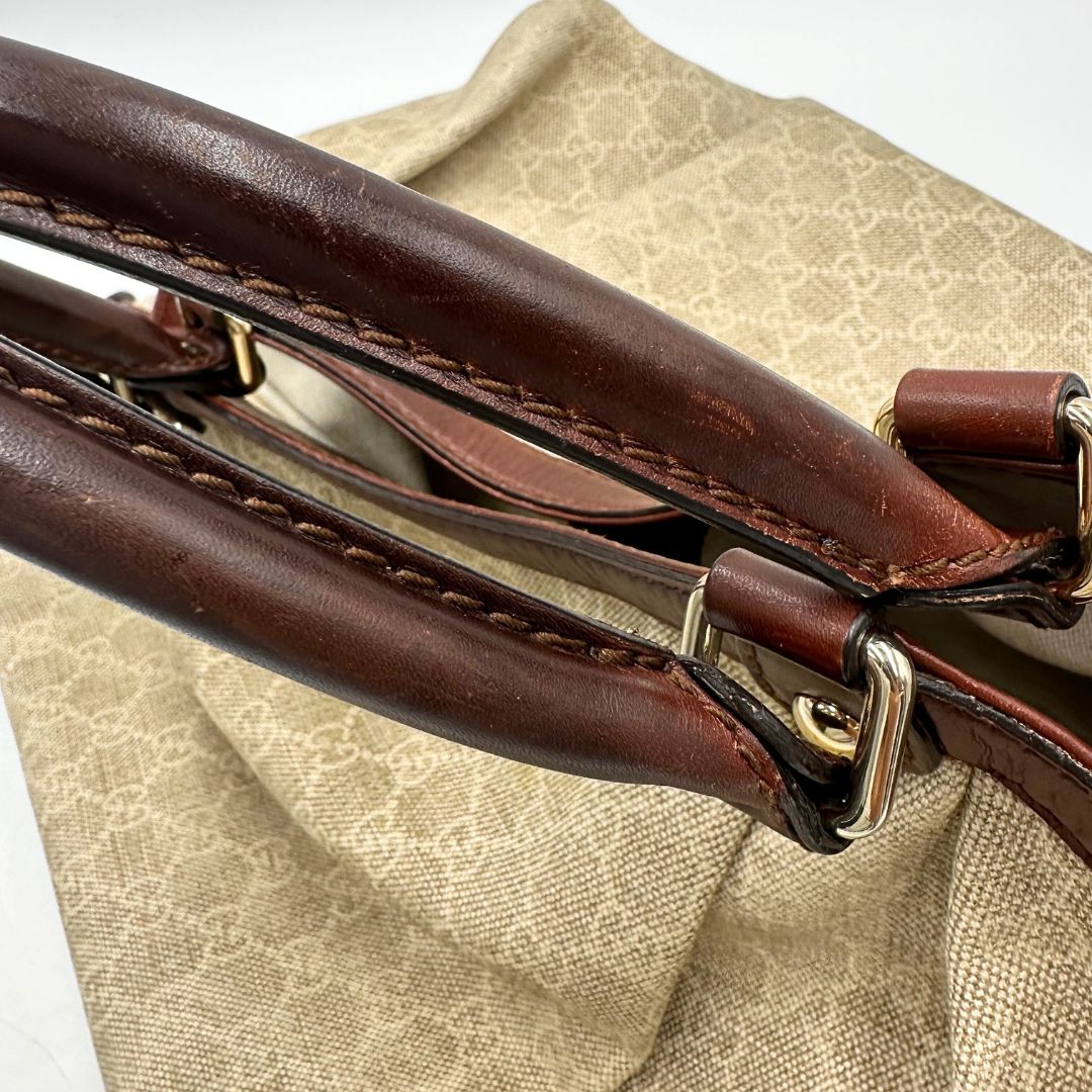 Manici di una borsa Gucci Sukey in tessuto beige GG con finiture in pelle marrone mogano, doppi manici stondati e ciondolo con doppie G incrociate. Rifinita con parti metalliche dorate. Indossabile a spalla.