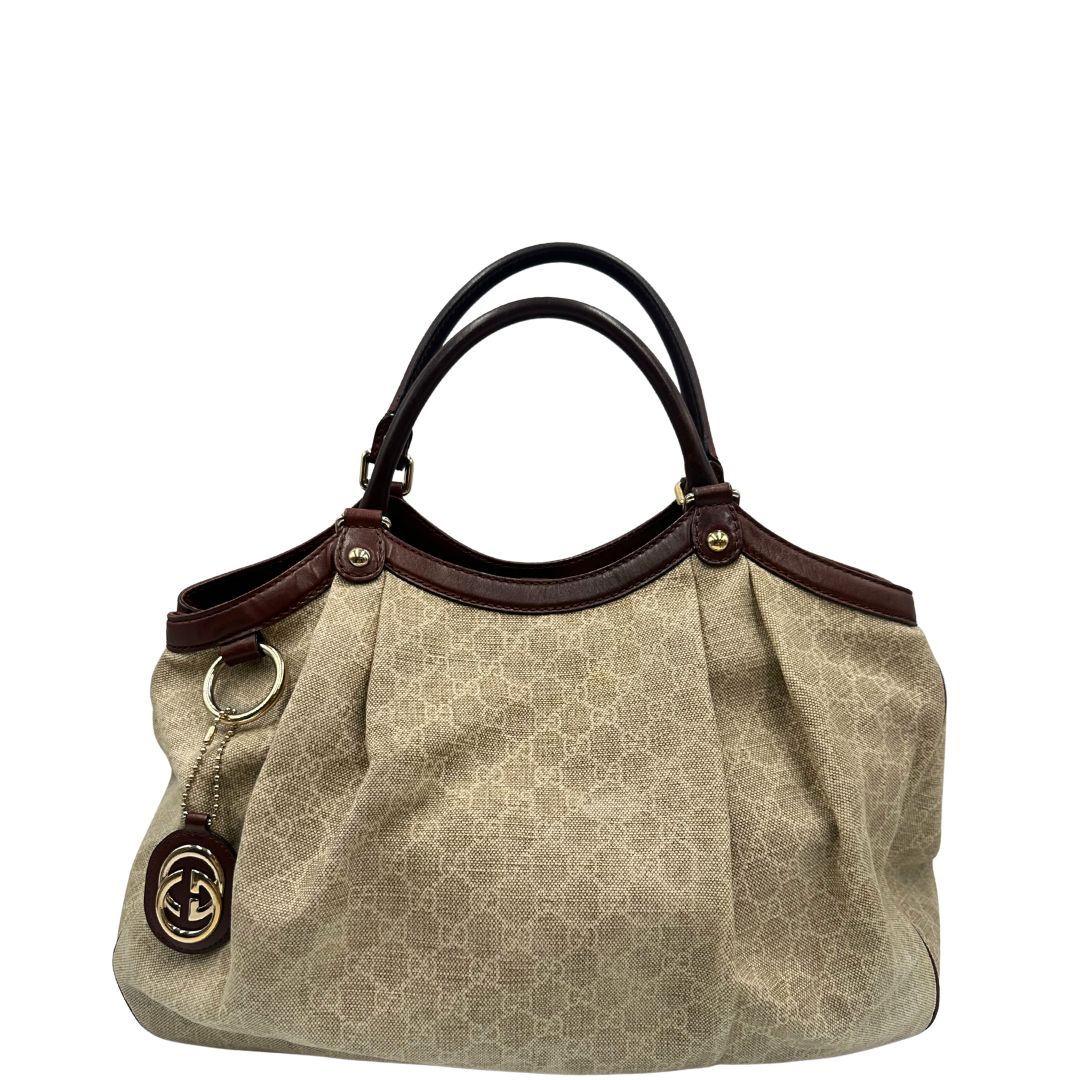 Front di una borsa Gucci Sukey in tessuto beige GG con finiture in pelle marrone mogano, doppi manici stondati e ciondolo con doppie G incrociate. Rifinita con parti metalliche dorate. Indossabile a spalla.