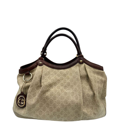 Front di una borsa Gucci Sukey in tessuto beige GG con finiture in pelle marrone mogano, doppi manici stondati e ciondolo con doppie G incrociate. Rifinita con parti metalliche dorate. Indossabile a spalla.