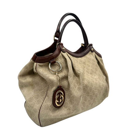 Laterale sinistro di una borsa Gucci Sukey in tessuto beige GG con finiture in pelle marrone mogano, doppi manici stondati e ciondolo con doppie G incrociate. Rifinita con parti metalliche dorate. Indossabile a spalla.