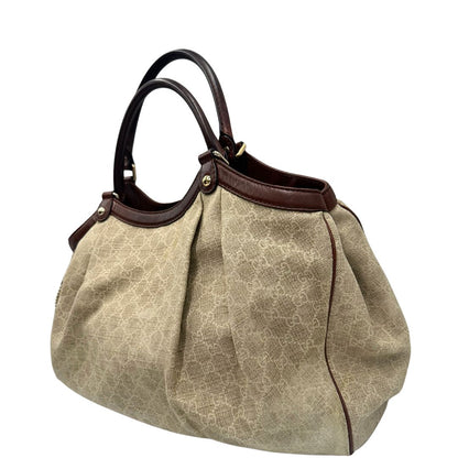 Laterale destro di una borsa Gucci Sukey in tessuto beige GG con finiture in pelle marrone mogano, doppi manici stondati e ciondolo con doppie G incrociate. Rifinita con parti metalliche dorate. Indossabile a spalla.
