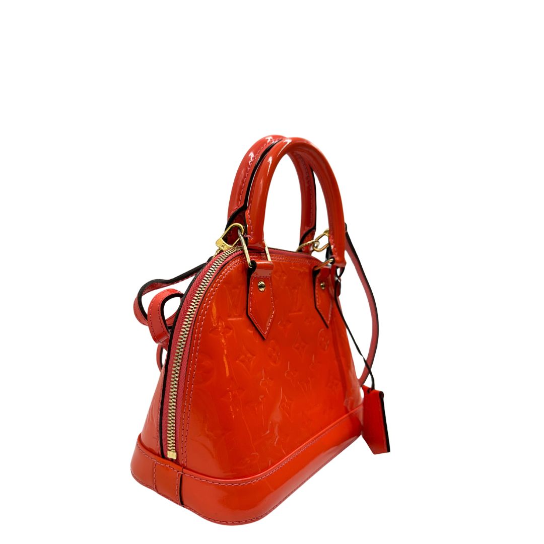 lato Borsa Louis Vuitton Alma BB in pelle Vernis arancione corallo monogram e parti metalliche dorate. Munita di doppi manici stondati e una tracolla amovibile, dotata di clochette porta chiavi. Completa di dustbag, lucchetto e chiavi, di lusso, originale, ottime condizioni, usata. 