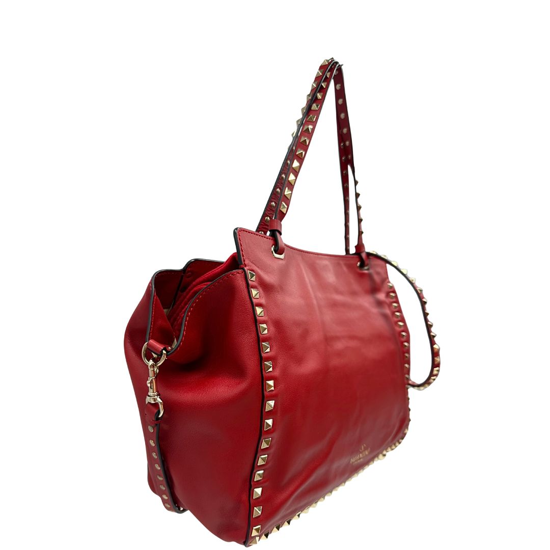 Laterale sinistro di una borsa Valentino Garavani in pelle rossa con parti metalliche dorate; impreziosita da borchie piramidali e munita di doppi manici e tracolla amovibile. Completa di dustbag. 