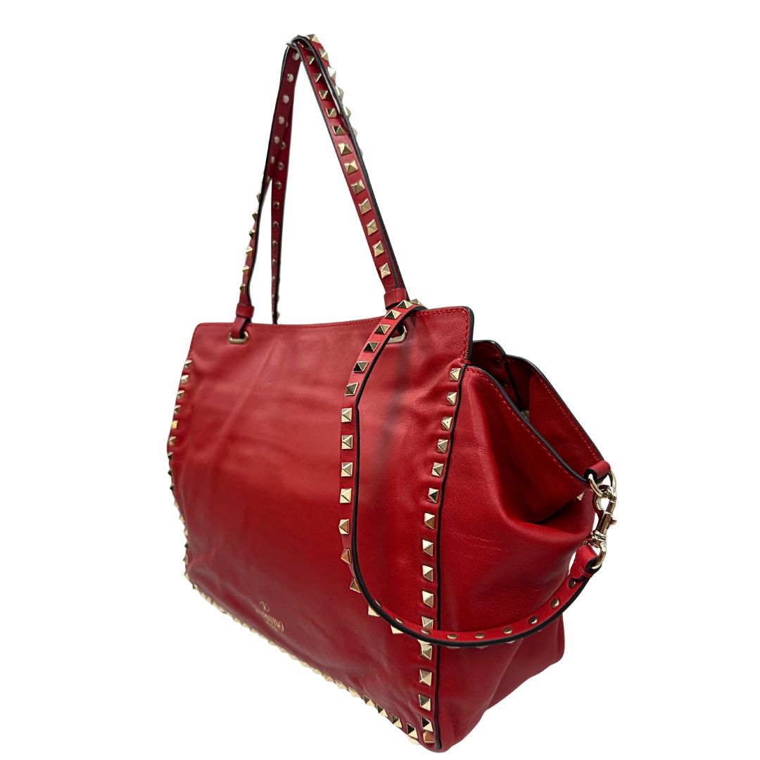 Laterale destro di una borsa Valentino Garavani in pelle rossa con parti metalliche dorate; impreziosita da borchie piramidali e munita di doppi manici e tracolla amovibile. Completa di dustbag. 