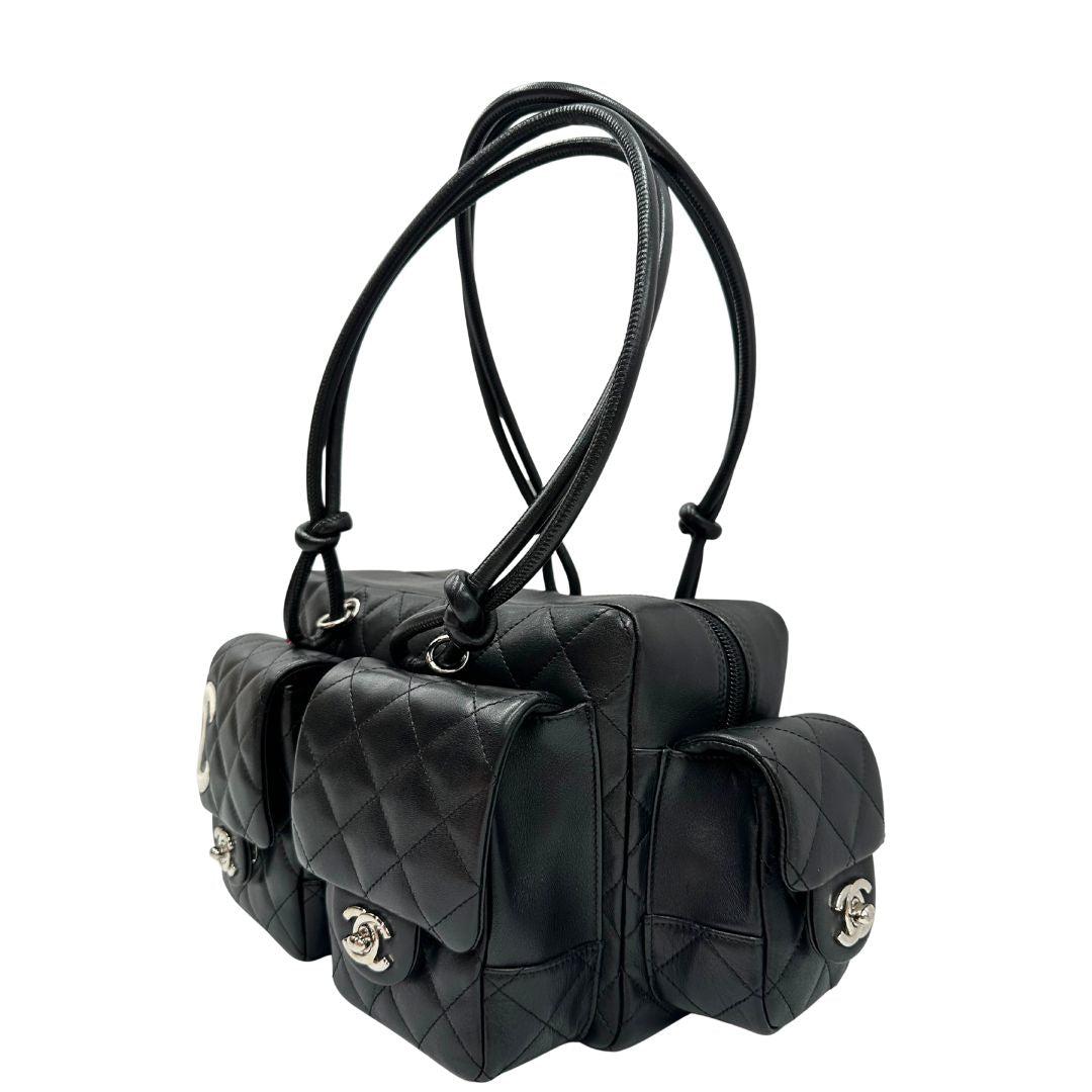 Laterale destro di una borsa Cambon Reporter Chanel in pelle trapuntata nera e doppie C bianche sul front, con tasche per accessori e doppi manici medio lunghi, rifinita con parti metalliche argentate. Completa di dustbag e card d'autenticità. Indossabile a spalla.