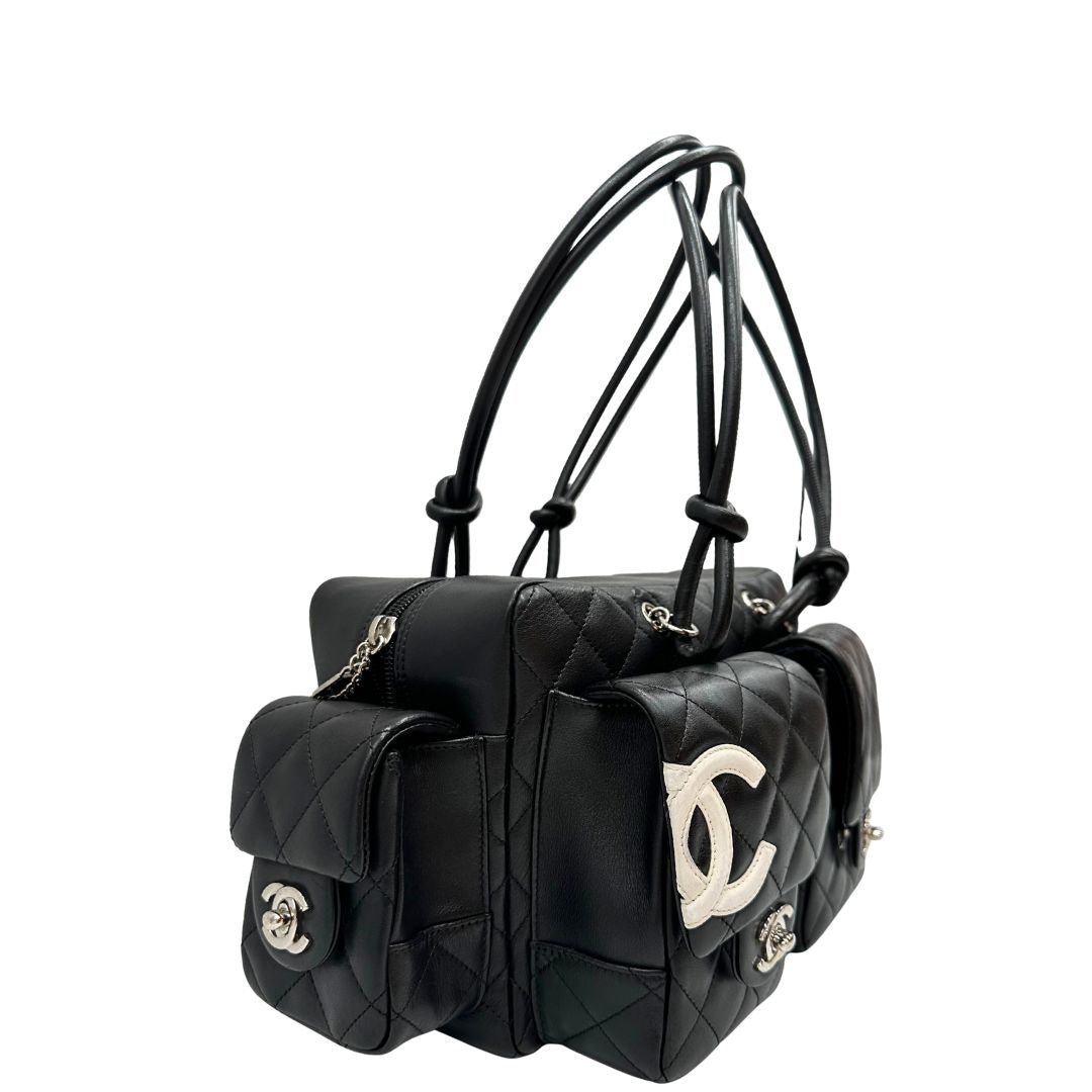 Laterale sinistro di una borsa Cambon Reporter Chanel in pelle trapuntata nera e doppie C bianche sul front, con tasche per accessori e doppi manici medio lunghi, rifinita con parti metalliche argentate. Completa di dustbag e card d'autenticità. Indossabile a spalla.