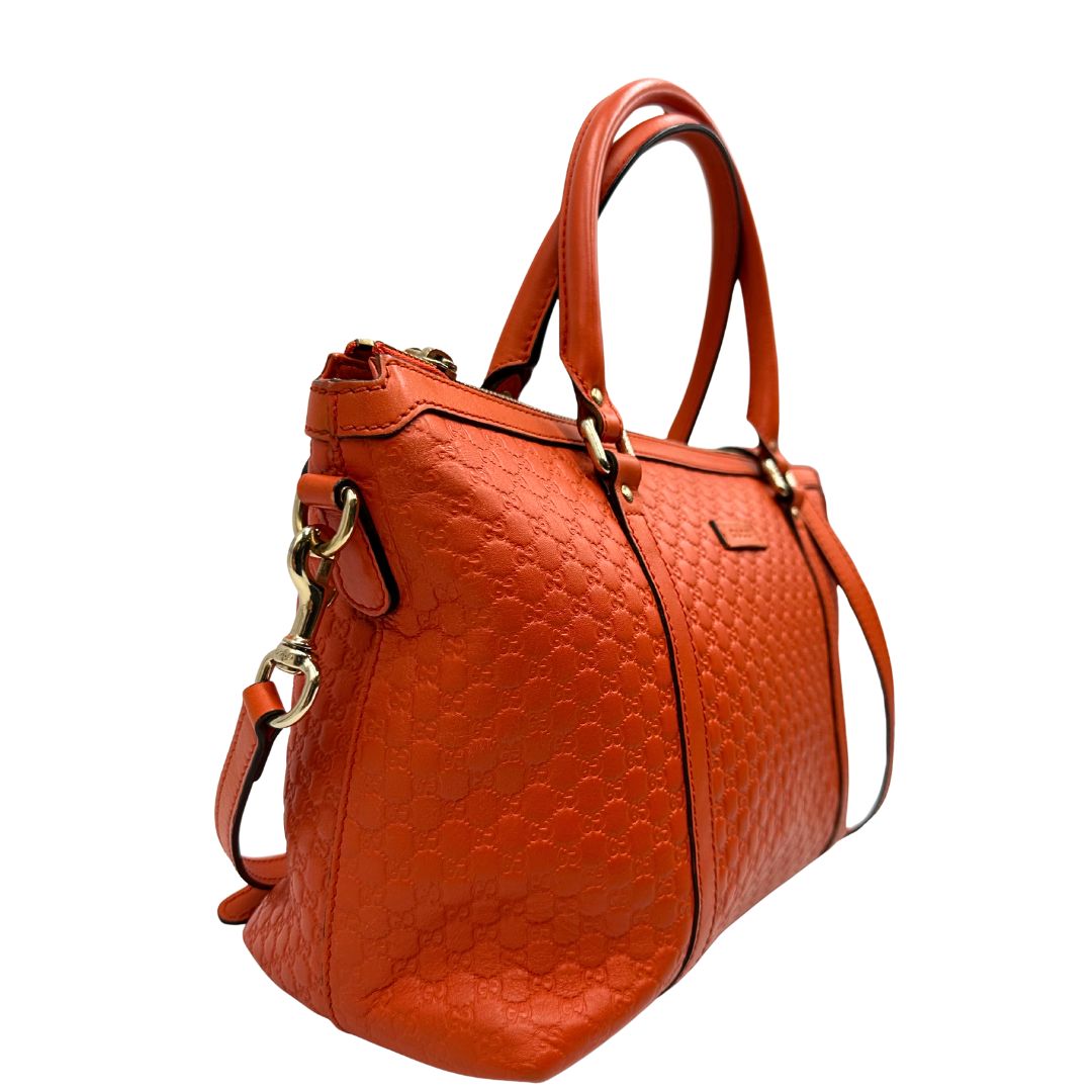 Laterale sinistro di una borsa Gucci in pelle arancione GG con parti metalliche dorate; munita di doppi manici e una tracolla amovibile e regolabile.