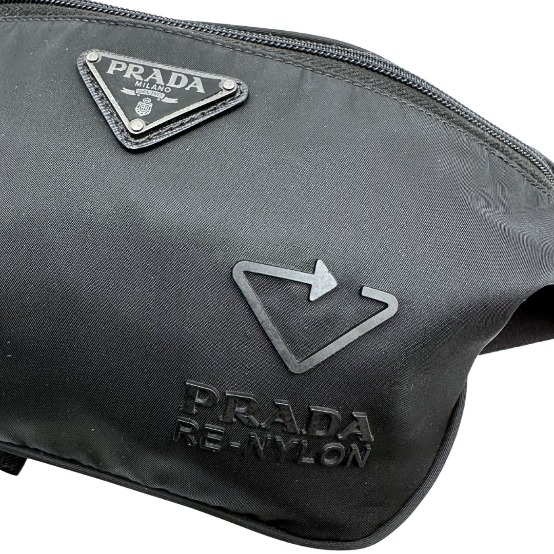 Loghi di un marsupio Prada in Re-Nylon nero con dettagli in pelle tono su tono, logo gommato sul front e parti in metallo argentato. Leggermente regolabile, indossabile a marsupio o cross body.