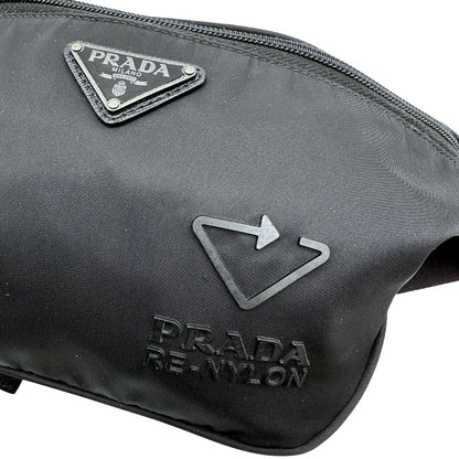 Loghi di un marsupio Prada in Re-Nylon nero con dettagli in pelle tono su tono, logo gommato sul front e parti in metallo argentato. Leggermente regolabile, indossabile a marsupio o cross body.