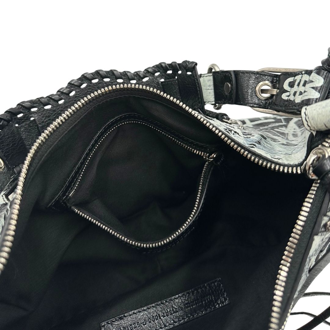 Interno di una borsa Balenciaga Le Cagole Graffiti in pelle nera con stampa graffiti color menta ghiacciata; dotata di manico leggermente regolabile con spallaccio. Pati metalliche argento anticato. Presenta tasca frontale con zip, lacci pendenti e ciondolo a cuore con specchietto integrato. Munita di scatola e dustbag. Indossabile a spalla.