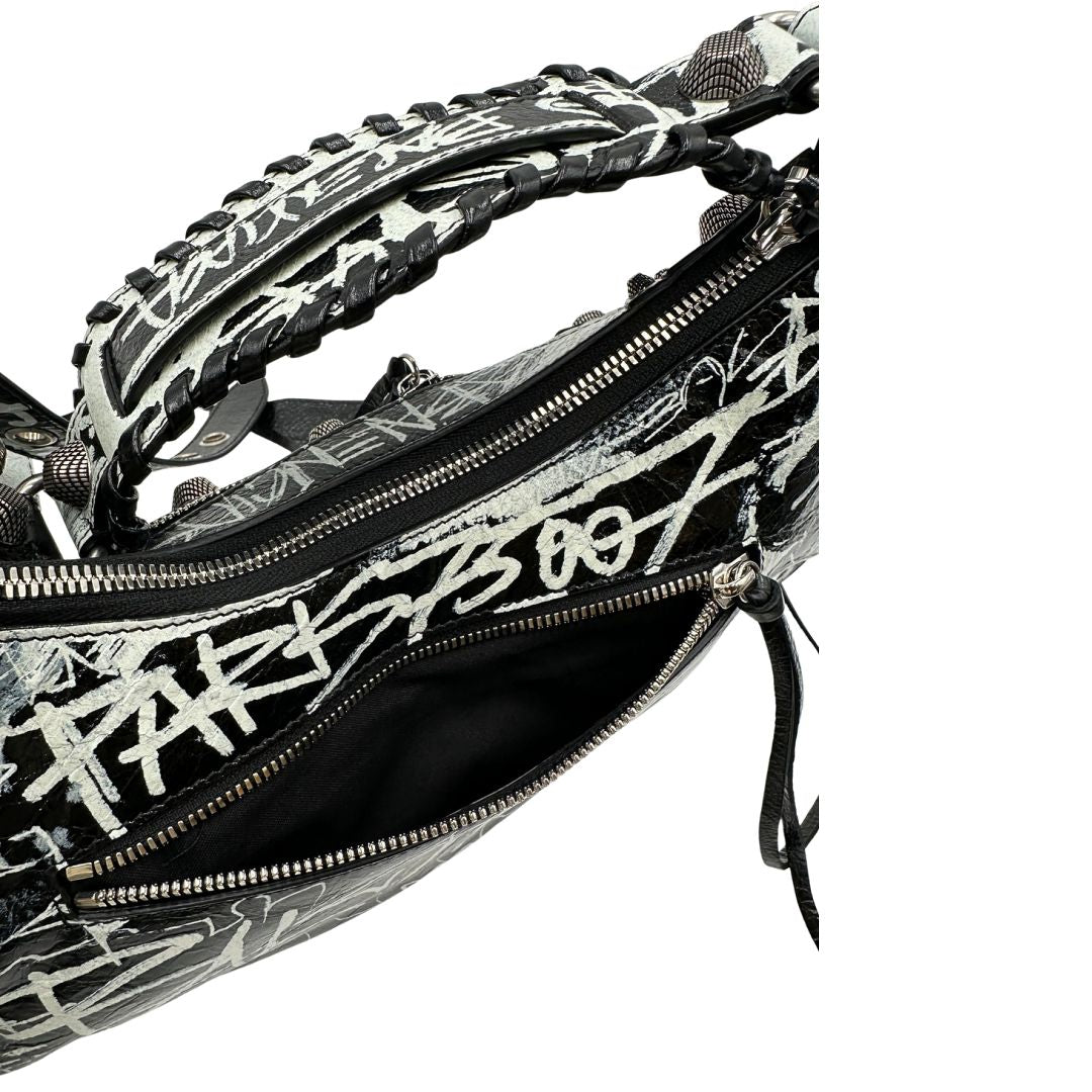 Vista superiore di una borsa Balenciaga Le Cagole Graffiti in pelle nera con stampa graffiti color menta ghiacciata; dotata di manico leggermente regolabile con spallaccio. Pati metalliche argento anticato. Presenta tasca frontale con zip, lacci pendenti e ciondolo a cuore con specchietto integrato. Munita di scatola e dustbag. Indossabile a spalla.