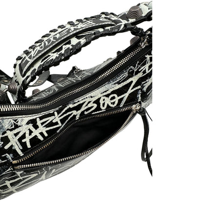 Vista superiore di una borsa Balenciaga Le Cagole Graffiti in pelle nera con stampa graffiti color menta ghiacciata; dotata di manico leggermente regolabile con spallaccio. Pati metalliche argento anticato. Presenta tasca frontale con zip, lacci pendenti e ciondolo a cuore con specchietto integrato. Munita di scatola e dustbag. Indossabile a spalla.
