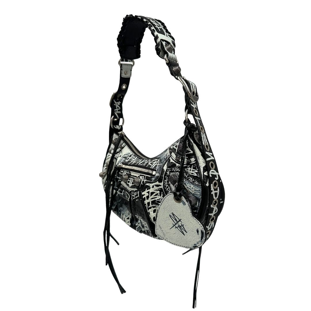 Laterale destro di una borsa Balenciaga Le Cagole Graffiti in pelle nera con stampa graffiti color menta ghiacciata; dotata di manico leggermente regolabile con spallaccio. Pati metalliche argento anticato. Presenta tasca frontale con zip, lacci pendenti e ciondolo a cuore con specchietto integrato. Munita di scatola e dustbag. Indossabile a spalla.