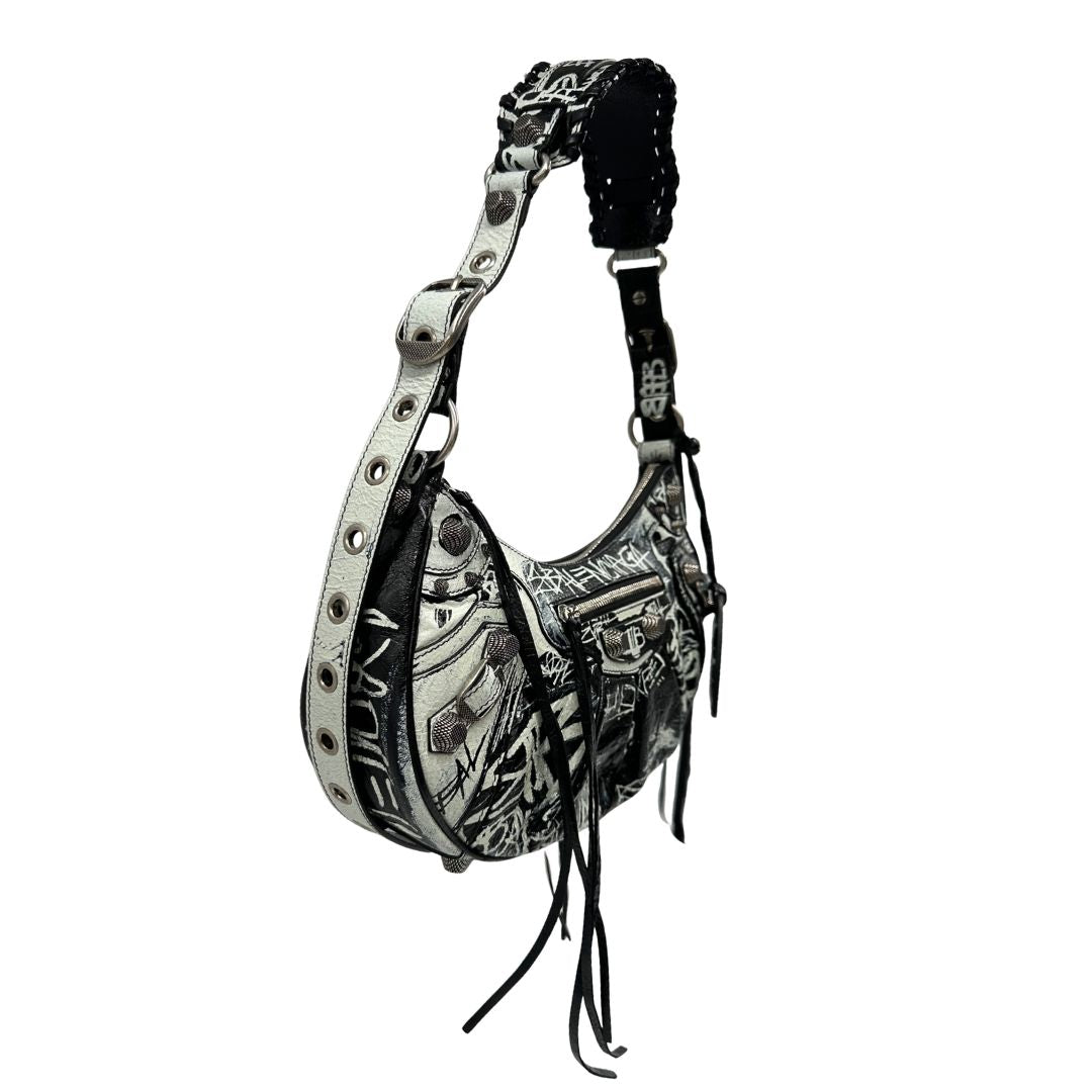 Laterale sinistro di una borsa Balenciaga Le Cagole Graffiti in pelle nera con stampa graffiti color menta ghiacciata; dotata di manico leggermente regolabile con spallaccio. Pati metalliche argento anticato. Presenta tasca frontale con zip, lacci pendenti e ciondolo a cuore con specchietto integrato. Munita di scatola e dustbag. Indossabile a spalla.