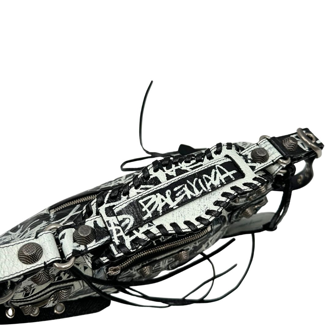 Vista superiore di una borsa Balenciaga Le Cagole Graffiti in pelle nera con stampa graffiti color menta ghiacciata; dotata di manico leggermente regolabile con spallaccio. Pati metalliche argento anticato. Presenta tasca frontale con zip, lacci pendenti e ciondolo a cuore con specchietto integrato. Munita di scatola e dustbag. Indossabile a spalla.