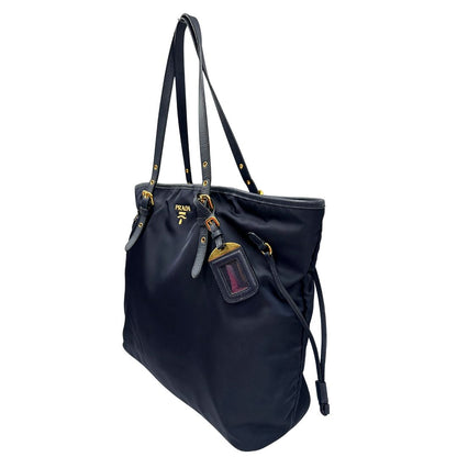 Laterale destro di una borsa Prada in nylon blu navy e finiture in pelle saffiano tono su tono, con parti metalliche dorate; munita di un accessorio porta address e doppi manici leggermente regolabili. Indossabile a spalla. Completa di dustbag e card d'autenticità.
