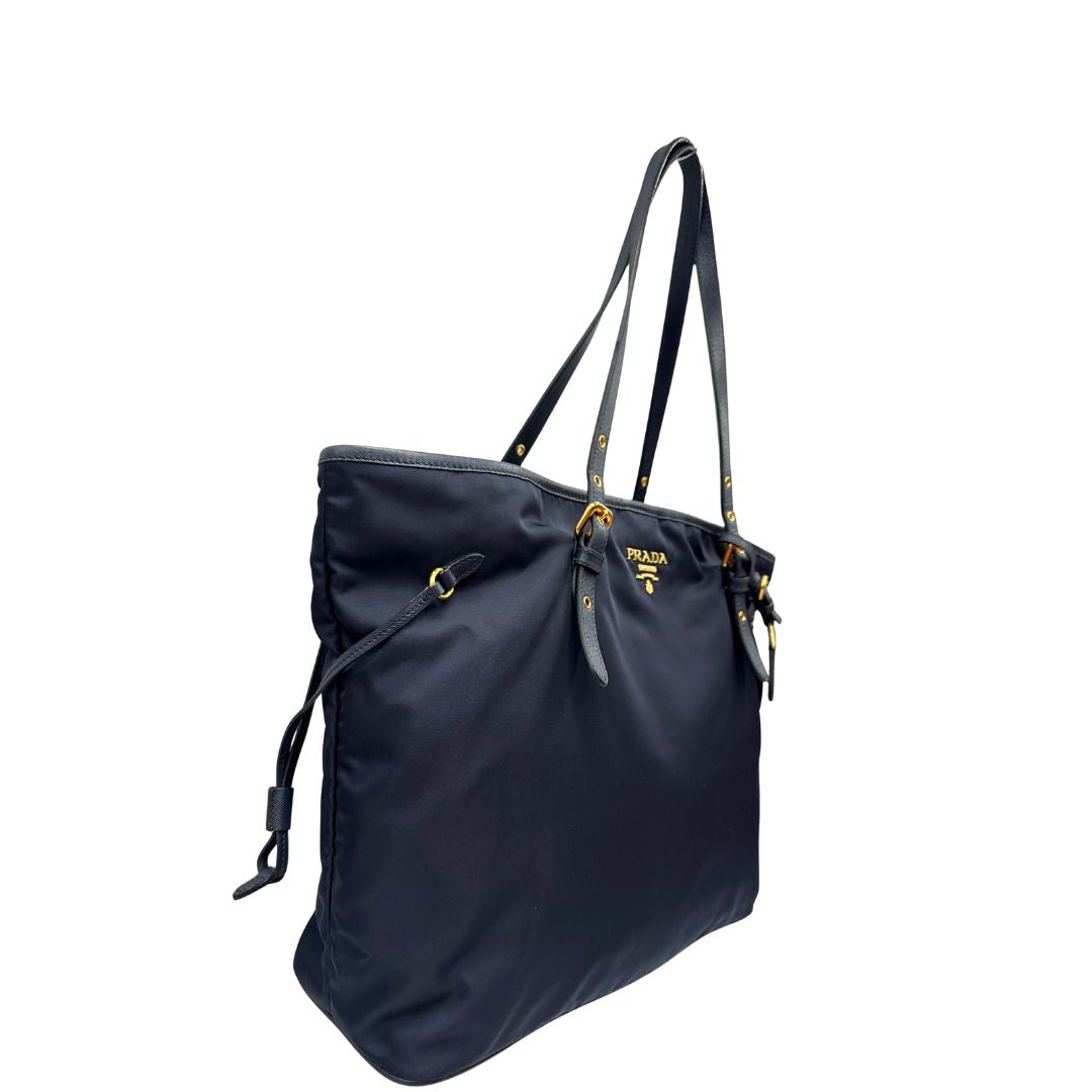 Laterale sinistro di una borsa Prada in nylon blu navy e finiture in pelle saffiano tono su tono, con parti metalliche dorate; munita di un accessorio porta address e doppi manici leggermente regolabili. Indossabile a spalla. Completa di dustbag e card d'autenticità.