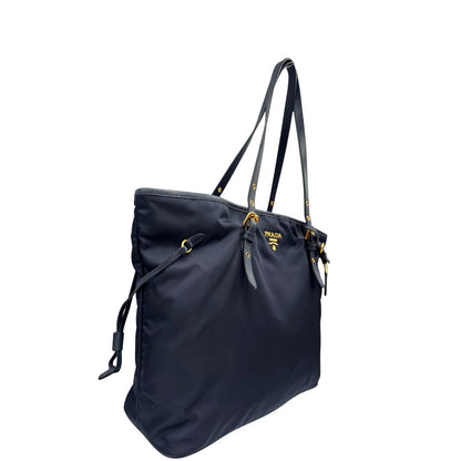 Laterale sinistro di una borsa Prada in nylon blu navy e finiture in pelle saffiano tono su tono, con parti metalliche dorate; munita di un accessorio porta address e doppi manici leggermente regolabili. Indossabile a spalla. Completa di dustbag e card d'autenticità.