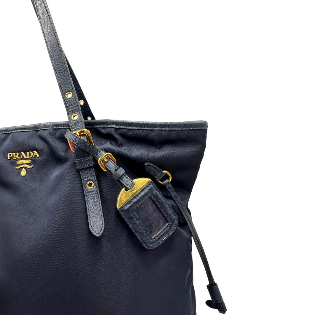 Tag porta address di una borsa Prada in nylon blu navy e finiture in pelle saffiano tono su tono, con parti metalliche dorate; munita doppi manici leggermente regolabili. Indossabile a spalla. Completa di dustbag e card d'autenticità.