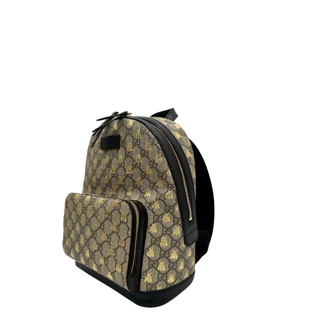Lato sinistro di un Zaino Gucci in canvas GG beige con finiture in pelle nere e parti metalliche dorate; presenta una stampa di api dorate su tutta la superficie, munito di un manico e bretelle in tessuto regolabili. Originale, usato, di lusso, in ottime condizioni.