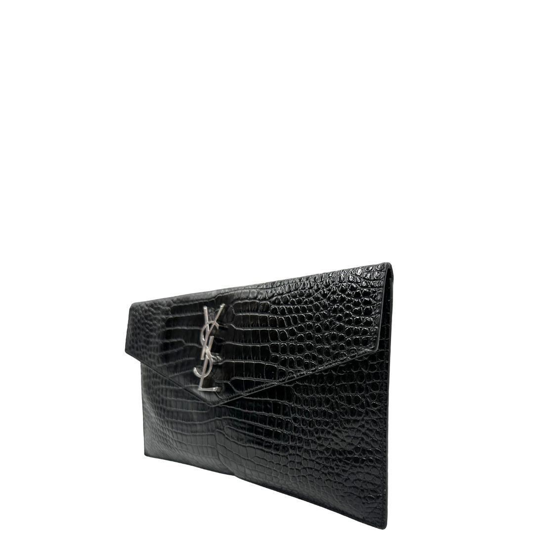 Laterale destro di una pochette Saint Laurent in pelle nera lucida effetto cocco, impreziosita dal logo Cassandre sul front in metallo argentato. Munita di dustbag.