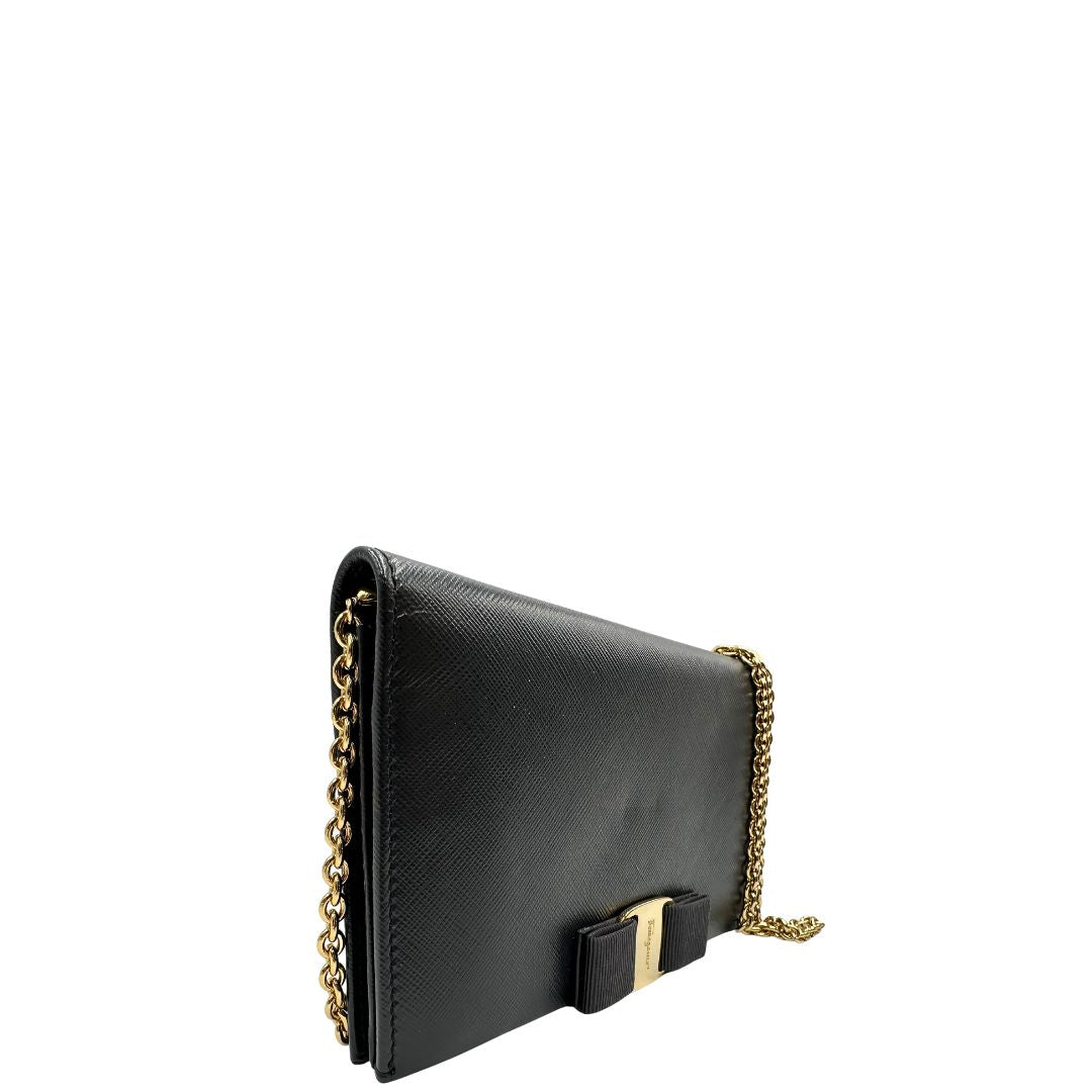 Laterale sinistro di un WOC Salvatore Ferragamo in pelle saffiano nera con parti metalliche oro. Munita di una tracolla in catena amovibile e non regolabile. Chiusura con fiocco. Completo di dustbag. Indossabile a tracolla.