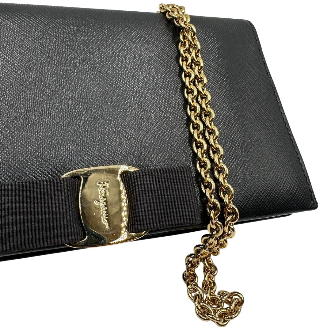 Fiocco della chiusura di un WOC Salvatore Ferragamo in pelle saffiano nera con parti metalliche oro. Munita di una tracolla in catena amovibile e non regolabile. Chiusura con fiocco. Completo di dustbag. Indossabile a tracolla.