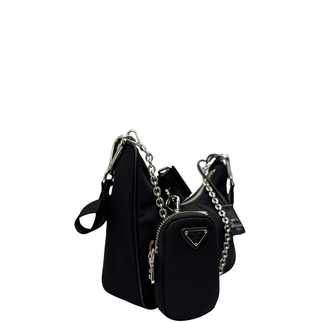 Laterale sinistro di una borsa Prada in nylon nero con parti metalliche argentate e finiture in pelle tono su tono; munita di una mini pouch amovibile, un accessorio portachiavi, manico in catena e una tracolla in tessuto amovibile e regolabile.