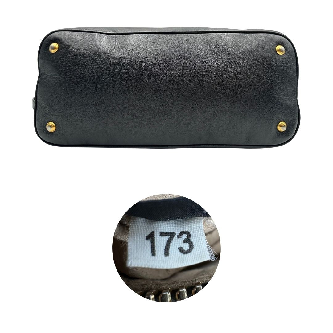Base e codice di una borsa Prada in pelle nera con parti metalliche dorate; munita di una tracolla amovibile e regolabile e doppi manici stondati.