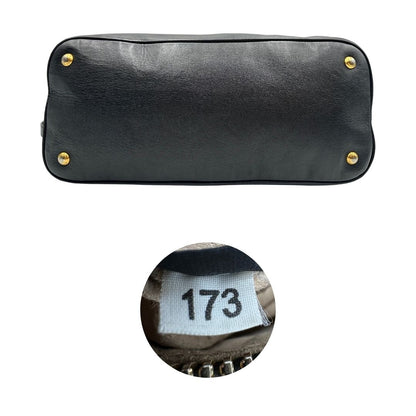 Base e codice di una borsa Prada in pelle nera con parti metalliche dorate; munita di una tracolla amovibile e regolabile e doppi manici stondati.