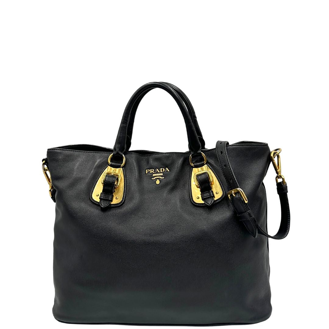 Front di una borsa Prada in pelle nera con parti metalliche dorate; munita di una tracolla amovibile e regolabile e doppi manici stondati.