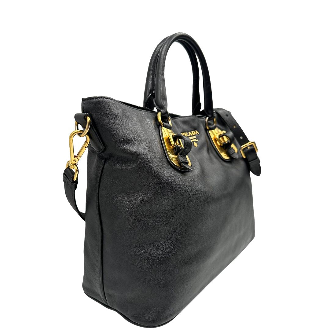 Laterale sinistro di una borsa Prada in pelle nera con parti metalliche dorate; munita di una tracolla amovibile e regolabile e doppi manici stondati.