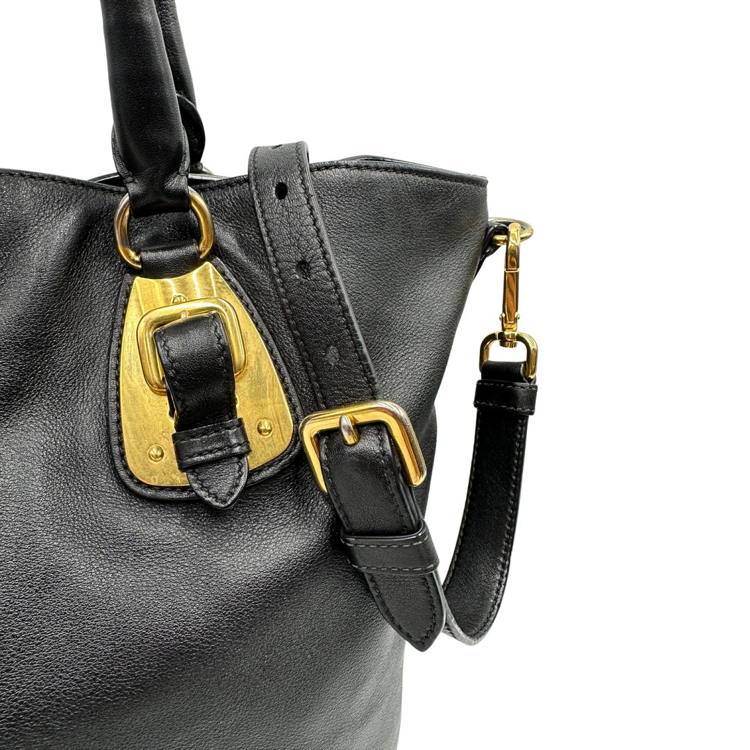 Fibbie di una borsa Prada in pelle nera con parti metalliche dorate; munita di una tracolla amovibile e regolabile e doppi manici stondati.