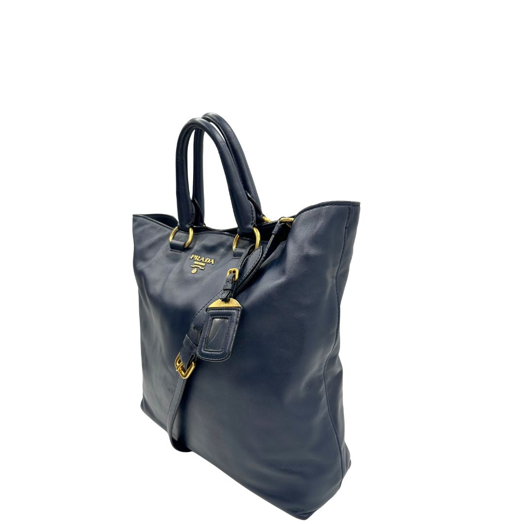 Lato sinistro di una Borsa Prada in pelle blu notte con parti metalliche dorate; munita di un accessorio porta address, doppi manici stondati e una tracolla amovibile e regolabile. Indossabile a mano oppure a tracolla. Originale, usata, di lusso, in ottime condizioni.