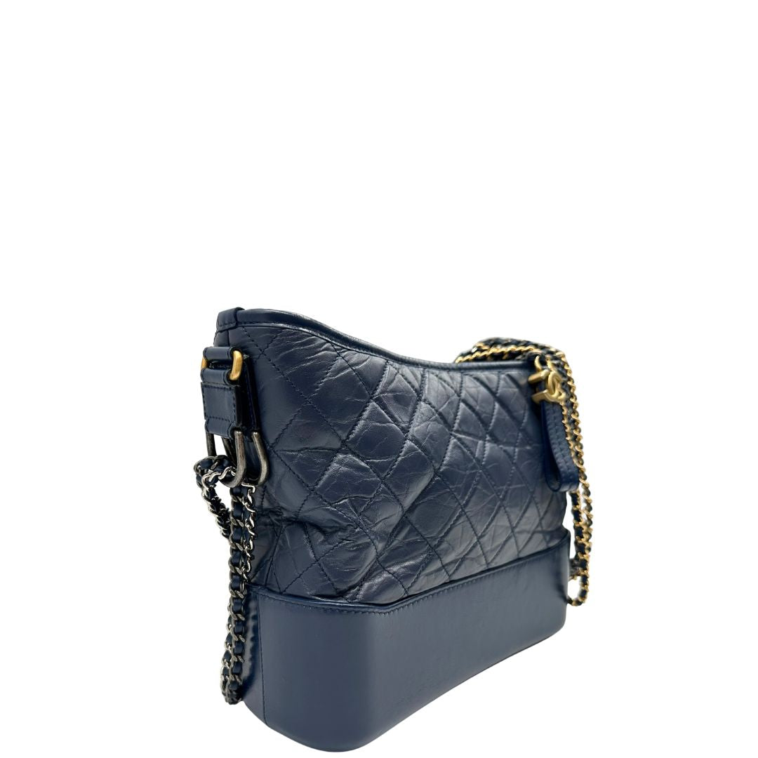 Laterale sinistro di una borsa Chanel Gabrielle in pelle matelassé blu navy con parti metalliche dorate e argentate. Munita di una tracolla in catena e pelle intrecciata con spallaccio. Tirante delle cerniera impreziosito dall'iconico logo CC in metallo dorato, completa di corredo.