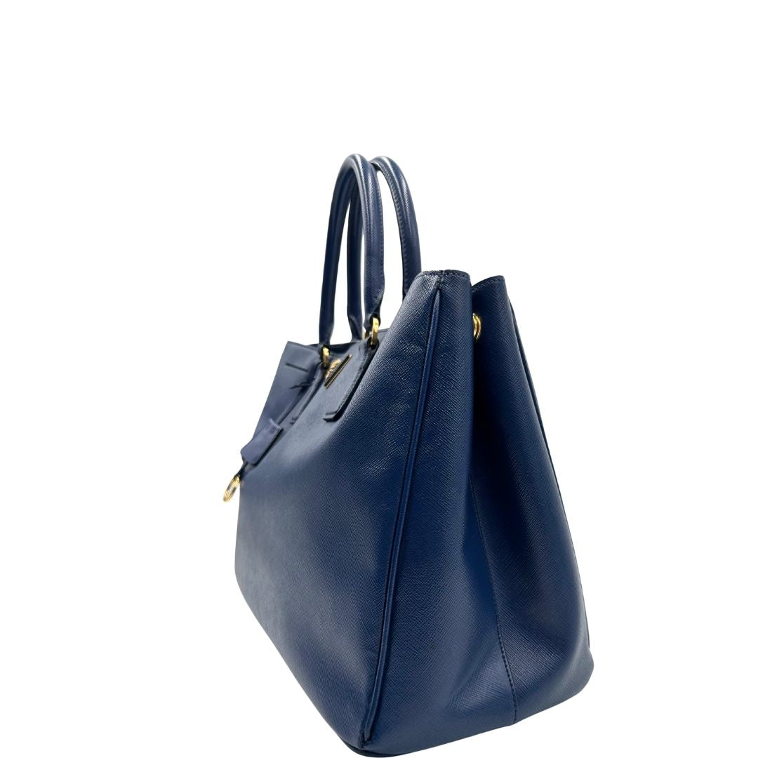 lato della Borsa Prada in pelle saffiano blu con parti metalliche dorate; munita di doppi manici stondati e impreziosita da una clochette portachiavi, di lusso, originale, ottime condizioni, usata.