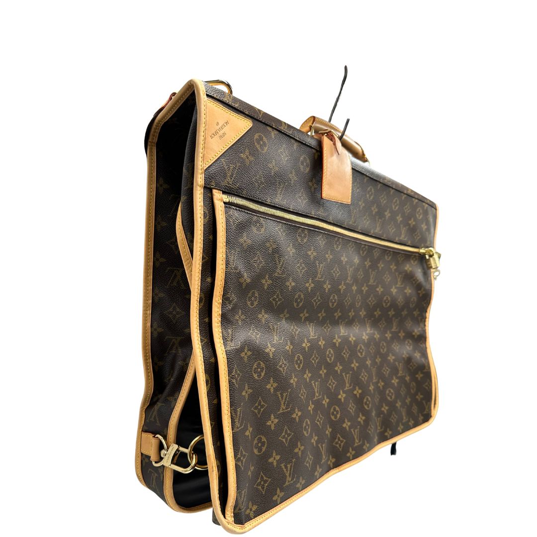 Lato destro di una Borsa da viaggio porta abiti in canvas marrone monogram con finiture in vacchetta naturale e parti metalliche dorate; munita di un manico singolo, una tracolla amovibile e regolabile e 2 grucce. Completa di Lucchetti e una chiave, dustbag.