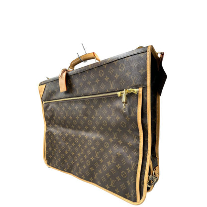 Lato sinistro di una Borsa da viaggio porta abiti in canvas marrone monogram con finiture in vacchetta naturale e parti metalliche dorate; munita di un manico singolo, una tracolla amovibile e regolabile e 2 grucce. Completa di Lucchetti e una chiave, dustbag.