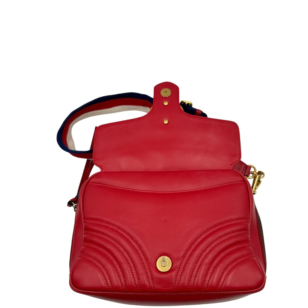 Apertura di una borsa Gucci Marmont in pelle matelassè rossa e parti metalliche dorate; munita di un manico superiore e di una tracolla in tessuto web blu/bianco/rosso, regolabile ed amovibile. Indossabile a mano o a tracolla. Completa di dustbag.