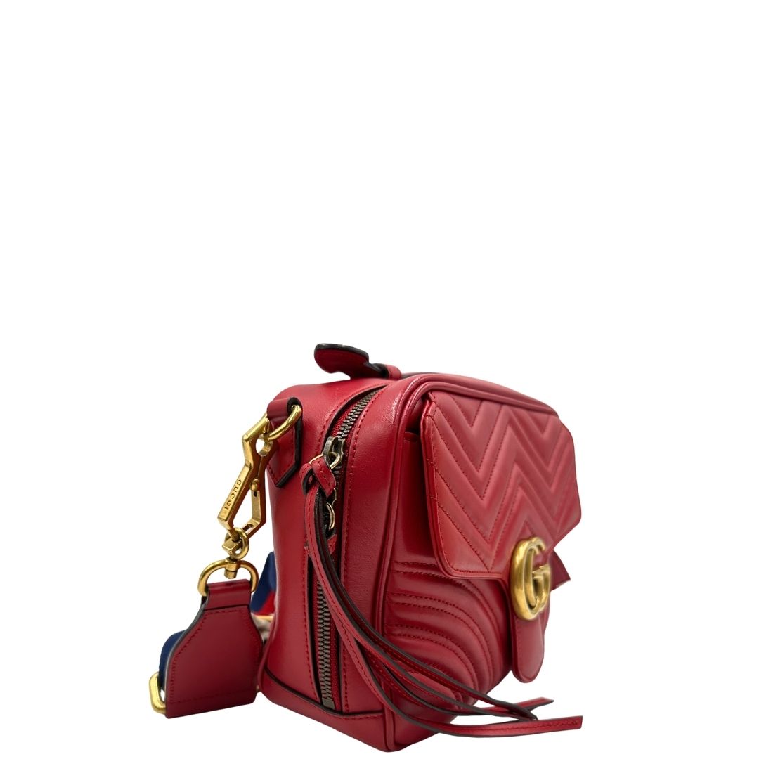 Laterale sinistro di una borsa Gucci Marmont in pelle matelassè rossa e parti metalliche dorate; munita di un manico superiore e di una tracolla in tessuto web blu/bianco/rosso, regolabile ed amovibile. Indossabile a mano o a tracolla. Completa di dustbag.