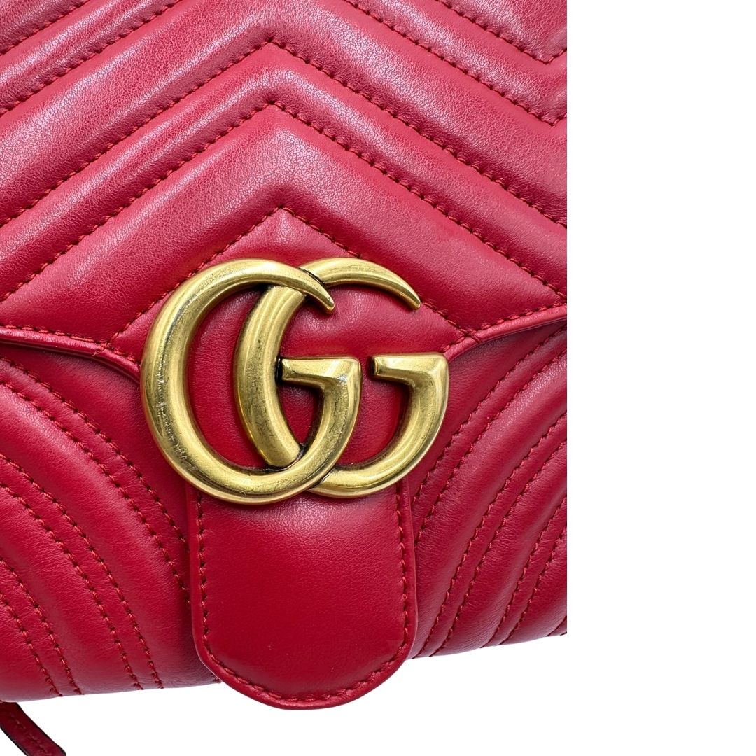 Dettaglio logo metallico di una borsa Gucci Marmont in pelle matelassè rossa e parti metalliche dorate; munita di un manico superiore e di una tracolla in tessuto web blu/bianco/rosso, regolabile ed amovibile. Indossabile a mano o a tracolla. Completa di dustbag.