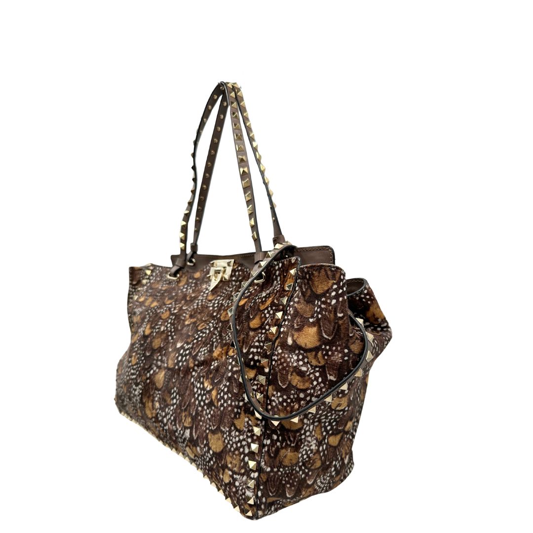 Lato sinstro di una Borsa Prada effetto cavallino stampato con piume multicolore e finiture in pelle marrone, con parti metalliche dorate; impreziosita da borchie piramidali e munita di doppi manici e tracolla amovibile. Completa di dustbag. Originale, usata, di lusso, in ottime condizioni.
