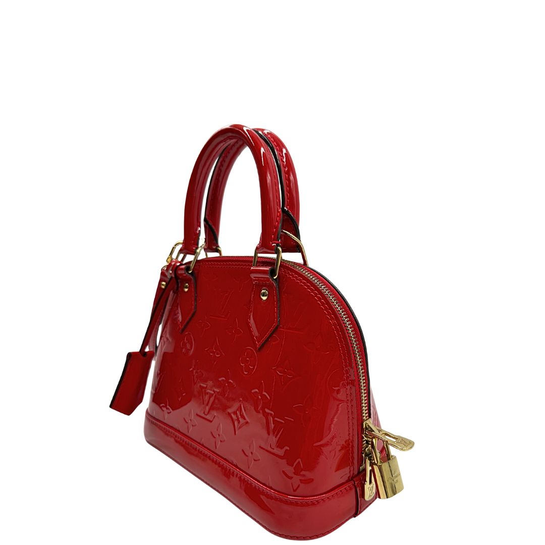 Laterale destro di una borsa Louis Vuitton Alma BB in pelle vernis rossa con trama monogram empreinte e parti metalliche dorate. Munita di doppi manici stondati e tracolla amovibile, dotata di clochette porta chiavi. Completa di scatola, dustbag, lucchetto e chiavi.