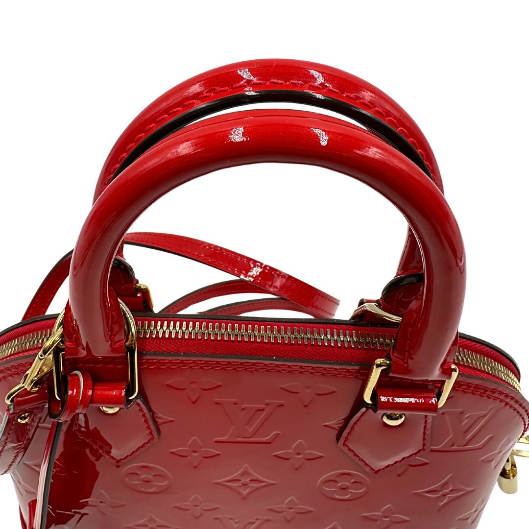 Manici di una borsa Louis Vuitton Alma BB in pelle vernis rossa con trama monogram empreinte e parti metalliche dorate. Munita di doppi manici stondati e tracolla amovibile, dotata di clochette porta chiavi. Completa di scatola, dustbag, lucchetto e chiavi.