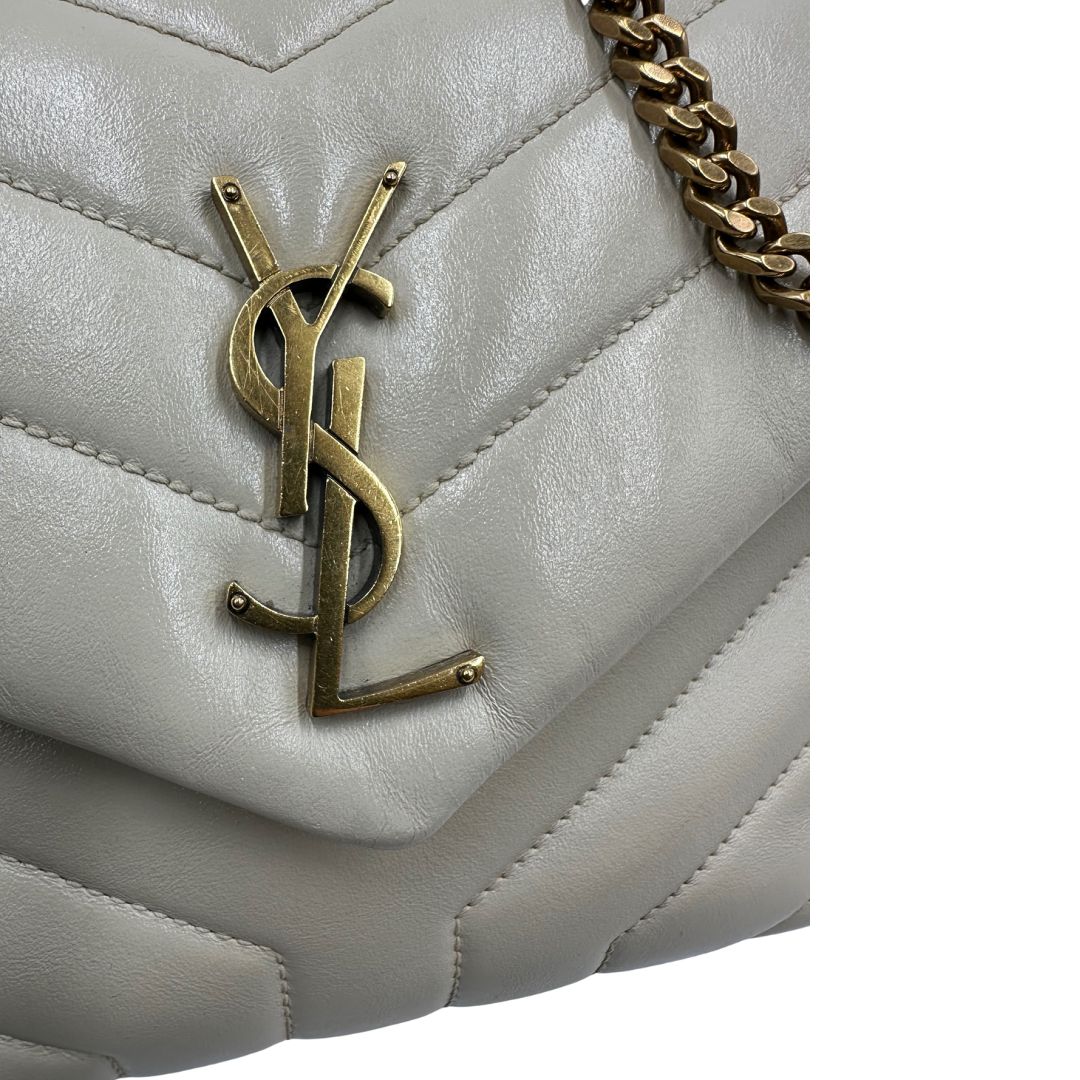Logo metallico di una borsa Saint Laurent in pelle trapuntata bianco latte con tracolla scorrevole in catena e spallacci in pelle, con parti metalliche dorate e iconico logo Cassandre sul front. Completa di dustbag. Indossabile a spalla o a tracolla.