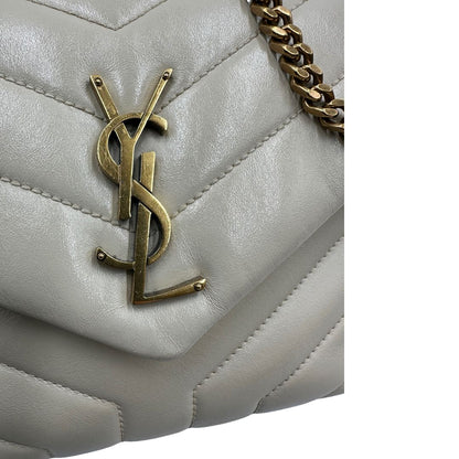 Logo metallico di una borsa Saint Laurent in pelle trapuntata bianco latte con tracolla scorrevole in catena e spallacci in pelle, con parti metalliche dorate e iconico logo Cassandre sul front. Completa di dustbag. Indossabile a spalla o a tracolla.