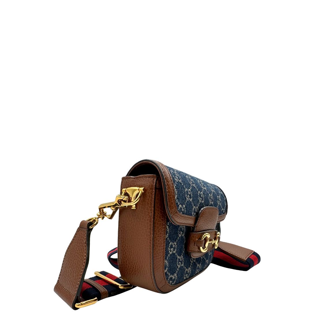 Laterale sinistro di una borsa Gucci in tessuto effetto denim con trama GG , finiture in pelle marroni e parti metalliche dorate; munita di una tracolla Web in tessuto blu e rossa e una tracolla in pelle, entrambe amovibili e regolabili. Completa di dustbag.