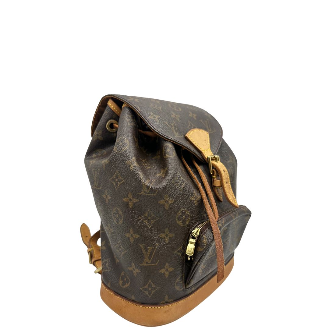 Laterale sinistro di uno zaino Montsouris mini in canvas marrone monogram con finiture in vacchetta naturale e parti metalliche dorate, munito di un anello sulla parte superiore e due bretelle in vacchetta regolabili con fibbie. Completo di dustbag.