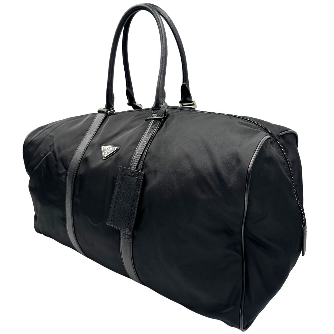 Laterale destro di un borsone Prada in nylon nero con finiture in pelle Saffiano tono su tono e parti metalliche argentate. Completo di dustbag, tag porta address e lucchetto. Indossabile a mano.