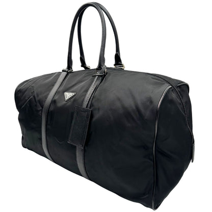 Laterale destro di un borsone Prada in nylon nero con finiture in pelle Saffiano tono su tono e parti metalliche argentate. Completo di dustbag, tag porta address e lucchetto. Indossabile a mano.